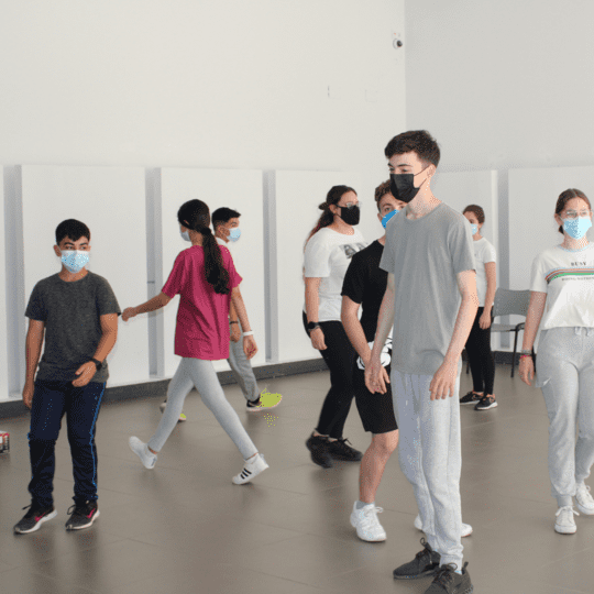 Niñas y niños en clases de teatro interactuando con mascarillas en un amplio salón.