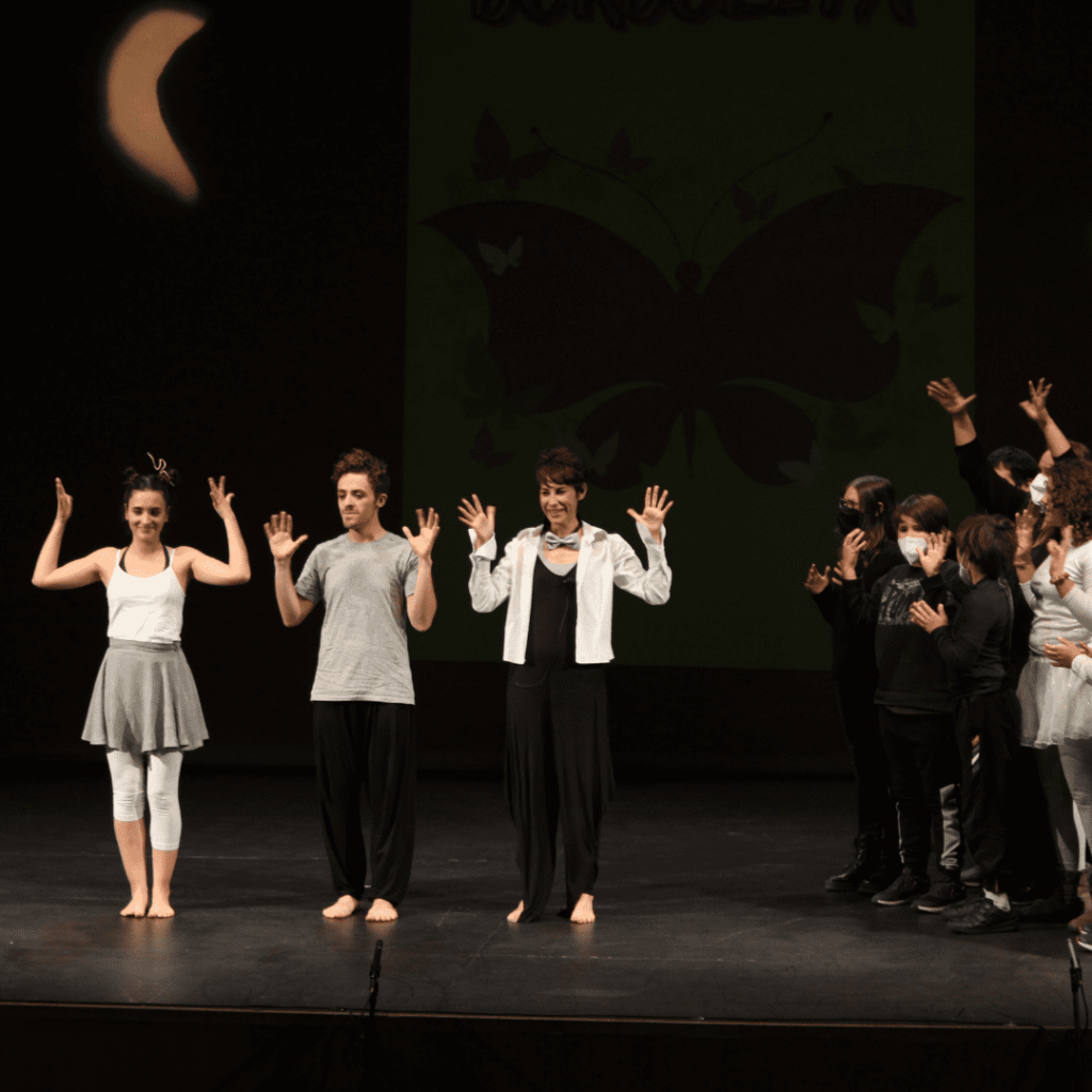 Personas adultas y niños sobre el escenario realizando una obra de teatro con un cartel de mariposa de fondo.